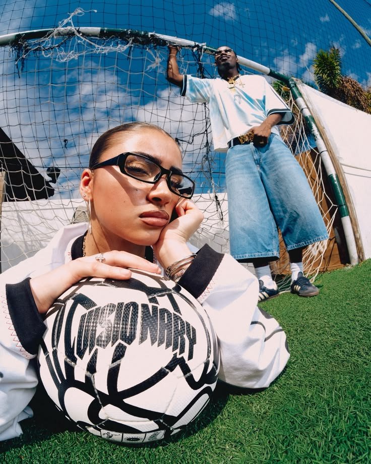 Two people in football-inspired outfits posing in front of a goal, one resting on a ball